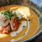Keto-friendly lobster bisque with saffron infusion, topped with butter-poached lobster, fresh microgreens, and a drizzle of coconut cream in a rustic ceramic bowl.