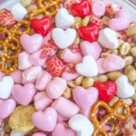 A colorful Cupid Crunch Valentine’s Day Trail Mix featuring heart-shaped candies, pink marshmallows, pretzels, peanuts, and dried strawberries in a glass bowl.