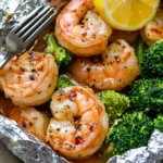 Close-up of baked shrimp and broccoli foil packs, featuring golden-browned shrimp with garlic butter seasoning, vibrant green broccoli, and a fresh lemon wedge.