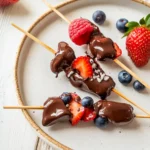 Keto chocolate berry skewers with strawberries, raspberries, and blueberries dipped in melted sugar-free chocolate on a ceramic plate.