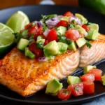 Keto Salmon with Avocado Salsa served on a black plate, featuring a crispy, pan-seared salmon fillet topped with chunky avocado salsa made of ripe avocados, cherry tomatoes, red onions, and cilantro. Garnished with fresh lime wedges.