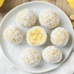 No-bake lemon and nut bliss balls coated in shredded coconut on a white plate, surrounded by lemons and macadamia nuts, viewed from above.