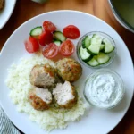 Juicy Greek chicken meatballs with cauliflower rice, cucumber-tomato salad, and creamy tzatziki sauce, served on a white plate with a gold fork on a wooden table.