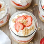 Layered strawberry cream delight in glass jars with whipped cream, fresh strawberries, and almond cookie crumbs on a wooden table.