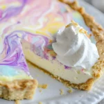 A beautifully swirled cheesecake pie with a pastel marbled top, almond flour crust, and a dollop of whipped cream, served on a white plate.