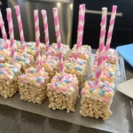 American Rice Krispie Treats squares with pastel sprinkles and pink striped sticks, set on parchment paper in a home kitchen.