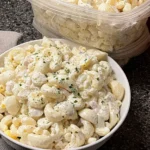 Creamy macaroni salad served in a white bowl, topped with parsley, with extra pasta salad in a plastic container behind it on a granite counter.