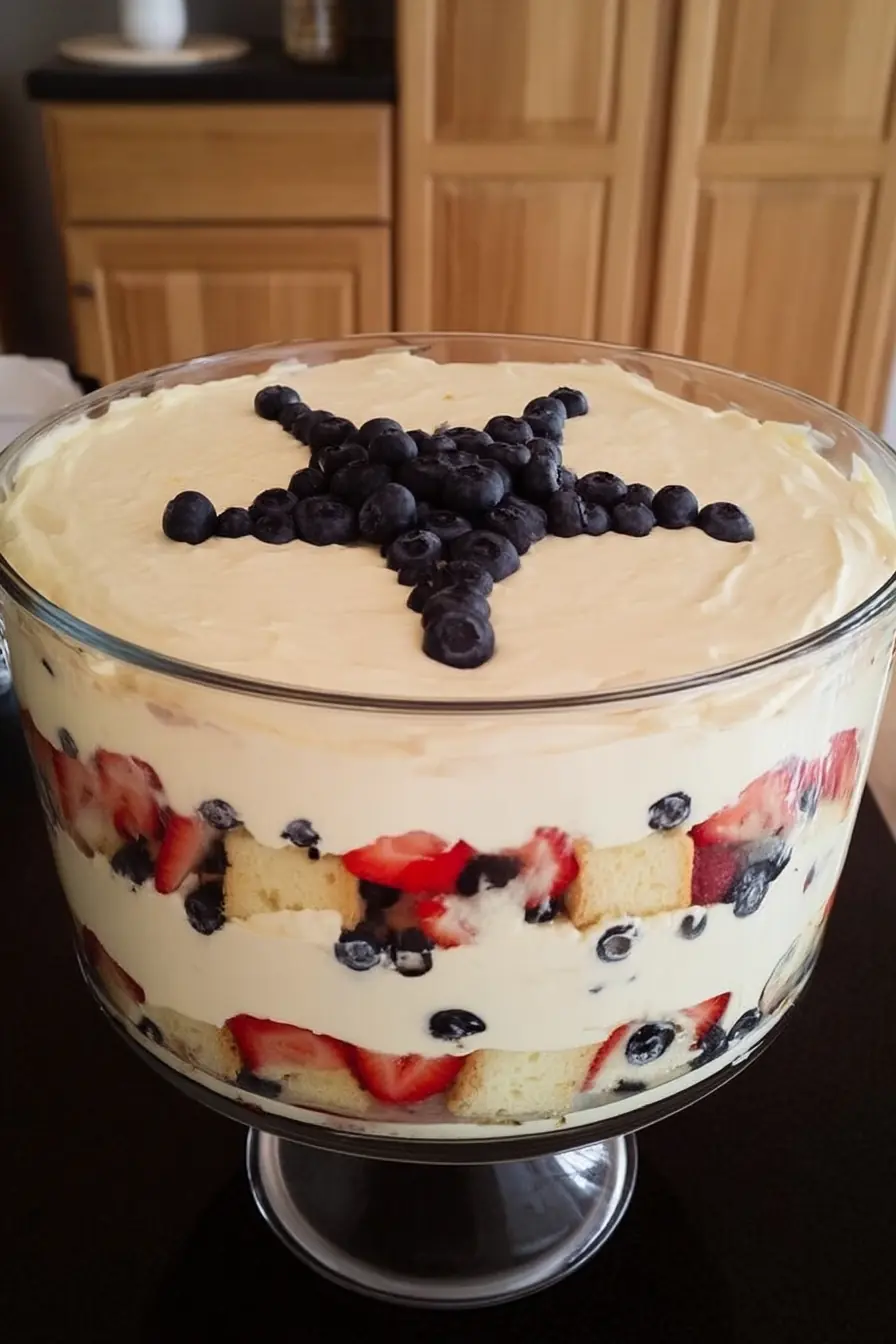 Layered 4th of July trifle in a clear glass bowl with visible strawberries, blueberries, pound cake, and pudding, topped with a star-shaped blueberry design.