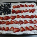 Patriotic Poke Cake topped with whipped cream, blueberries, and sliced strawberries in a flag design