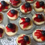 Patriotic mini cheesecakes topped with sliced strawberries and blueberries on a glass platter