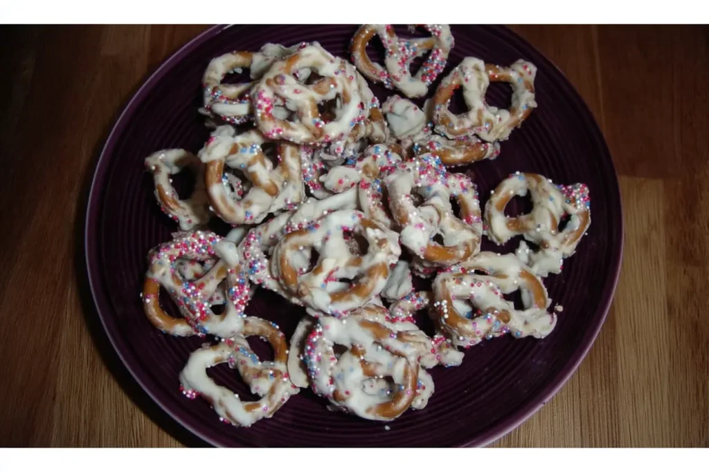 White chocolate-covered pretzels with red, white, and blue sprinkles on a dark plate – a festive patriotic snack mix idea.