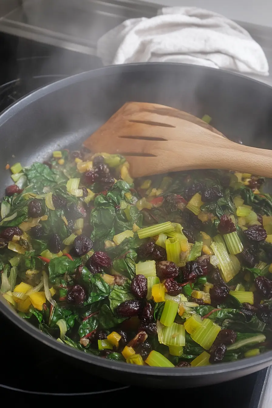 Sautéed Swiss chard in a skillet with golden stems, dried cranberries, and a wooden spatula stirring the greens