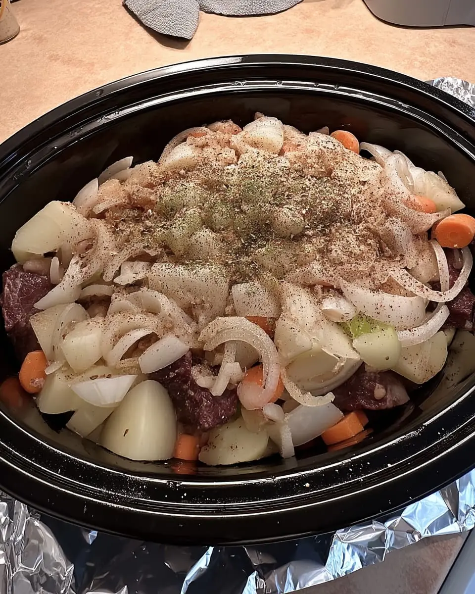 Slow cooker beef stew ingredients—cubed beef, potatoes, carrots, onions, and herbs—layered in a black crockpot before cooking.