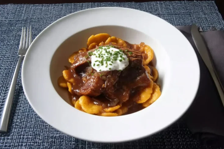 Overhead view of apple cider braised pork shoulder on golden spätzle, topped with crème fraîche and chopped chives in a white bowl.