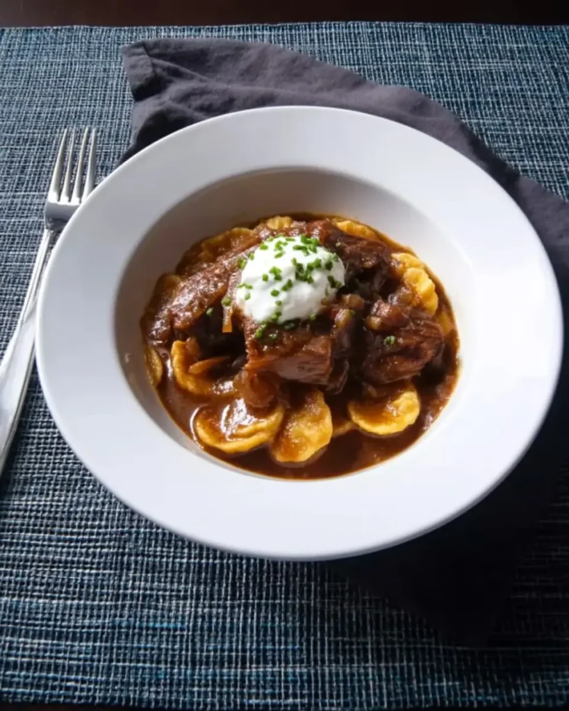 Apple cider braised pork shoulder in rich cider gravy over spätzle, topped with crème fraîche and fresh chives in a white bowl on a blue placemat.