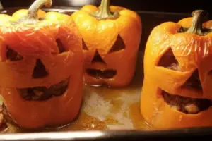 Three baked stuffed jack-o-lantern bell peppers with carved faces in baking dish with juices, showing tender peppers and cooked beef filling