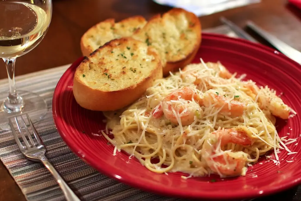 Shrimp scampi pasta with angel hair noodles, pink shrimp, and Parmesan cheese on red plate, served with toasted garlic bread and white wine