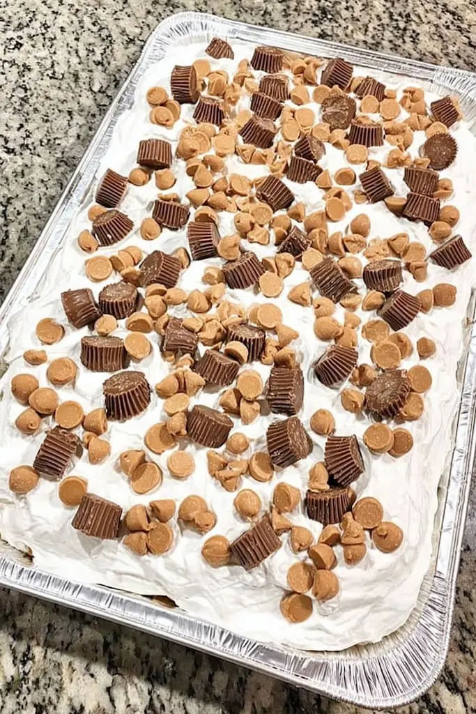 Full pan of Better Than Anything peanut butter poke cake in aluminum pan topped with whipped cream, mini Reese's peanut butter cups, and peanut butter chips on granite counter