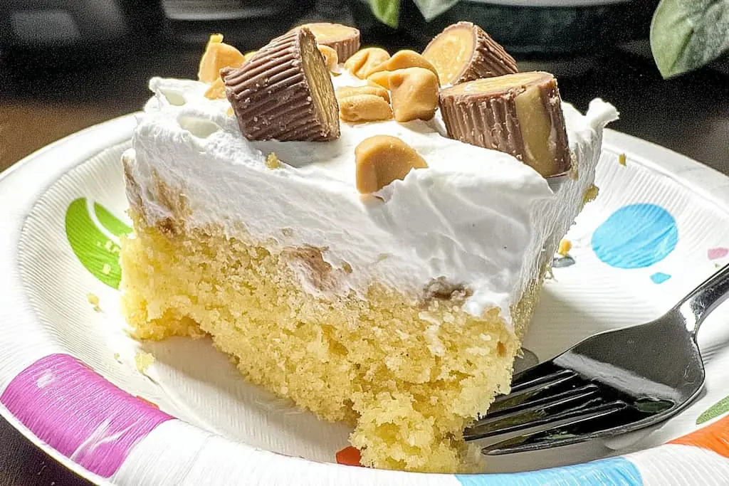 Slice of Better Than Anything peanut butter poke cake on colorful plate with fork showing moist yellow cake soaked with peanut butter filling and topped with whipped cream and mini Reese's cups
