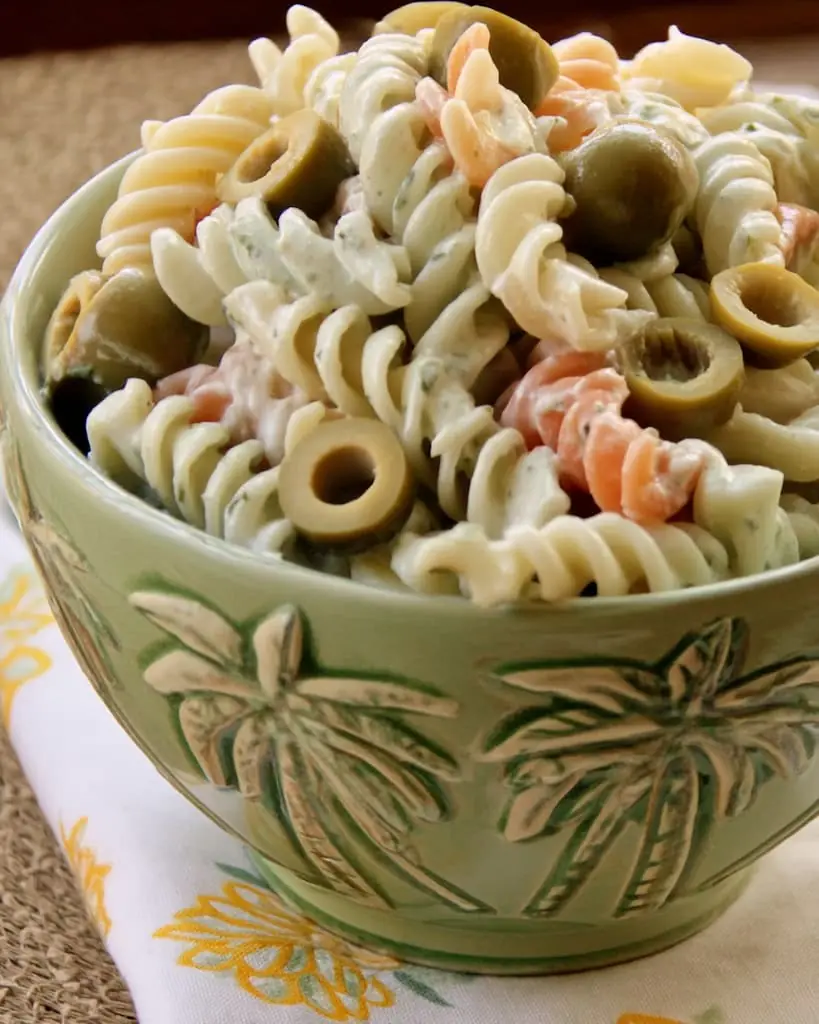 Close-up of tri-color rotini coated in creamy dill pickle dressing, topped with sliced green olives, served in a green ceramic bowl with palm tree pattern.