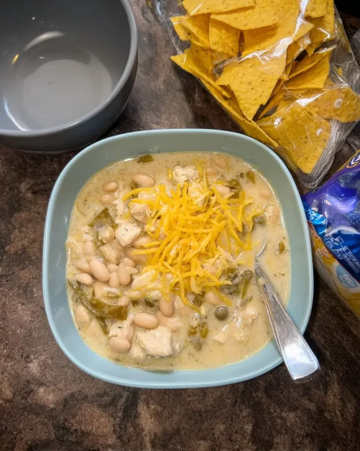 Bowl of creamy white chicken chili topped with shredded cheddar cheese, served with tortilla chips on wooden table