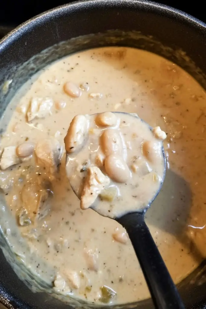 Ladle full of thick creamy white chicken chili with beans and chicken pieces cooking in dark pot