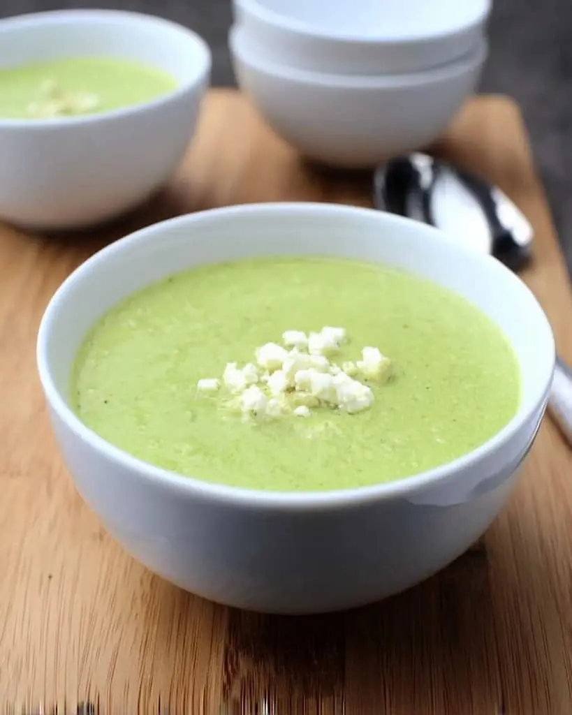 Creamy green zucchini soup in white bowl topped with crumbled feta cheese on wooden cutting board