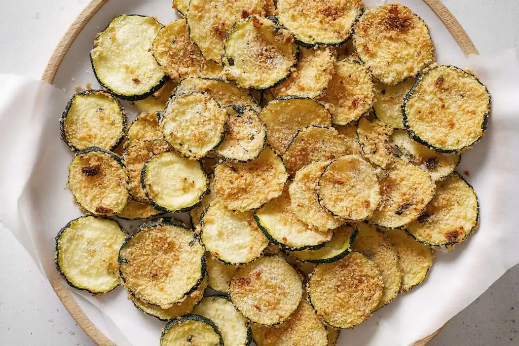 Golden crispy baked zucchini chips with breadcrumb coating spread on a white ceramic plate