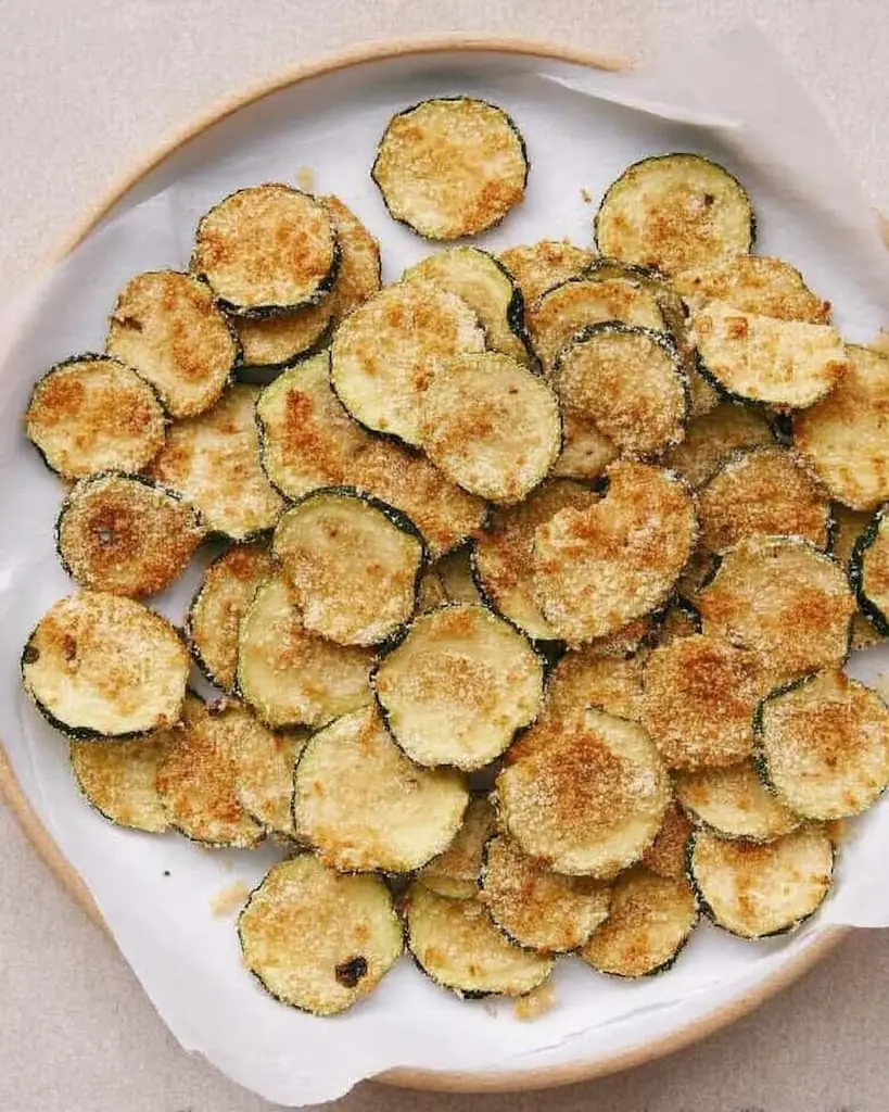 Golden brown crispy baked zucchini chips with Parmesan coating arranged on a white ceramic plate