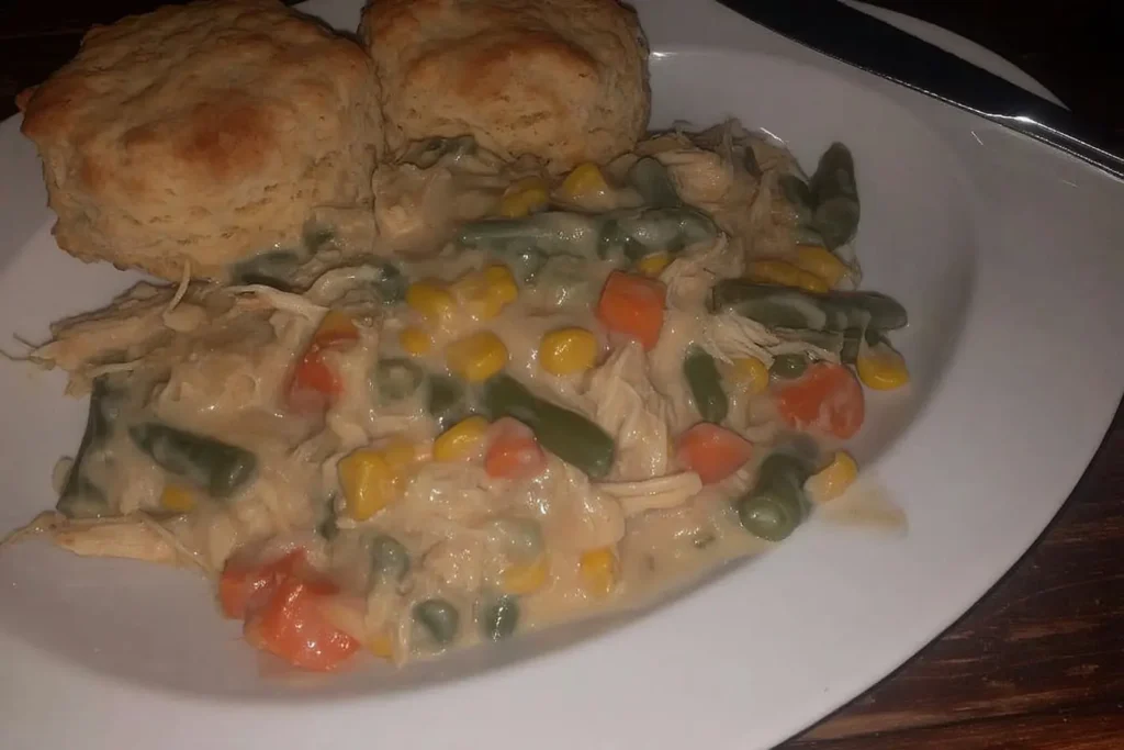 Creamy crockpot chicken pot pie filling with shredded chicken, carrots, corn, peas, and green beans, plated alongside two golden biscuits on a white dish.