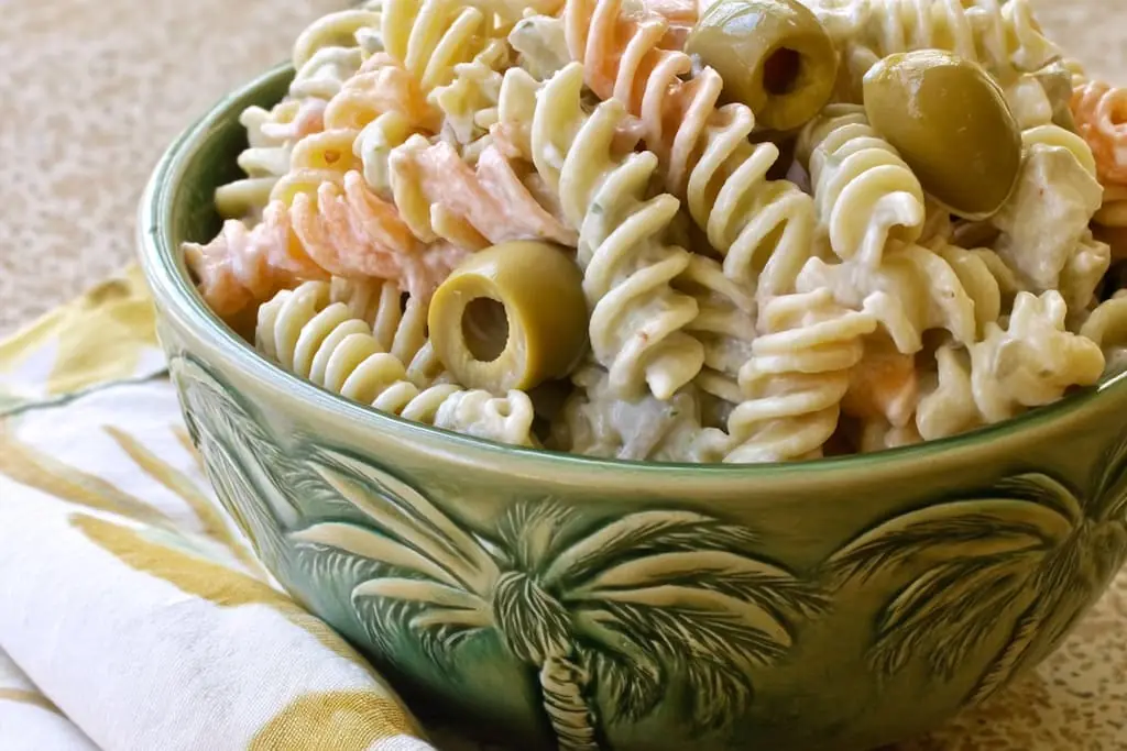 Tri-color rotini coated in creamy dill pickle dressing with sliced green olives, served in a green palm-pattern ceramic bowl on a yellow patterned napkin.
