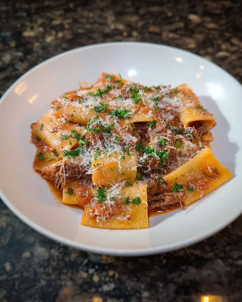 Pappardelle pasta with braised short rib ragu on a white plate, topped with grated Parmesan cheese and fresh parsley