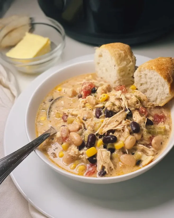 Creamy white chicken chili in a white bowl topped with shredded chicken, black beans, corn, and diced tomatoes, served with crusty bread and butter