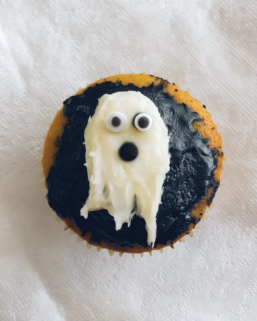 Halloween ghost cupcake with black frosting base, white ghost-shaped frosting dripping down sides, and candy eyes on white paper liner