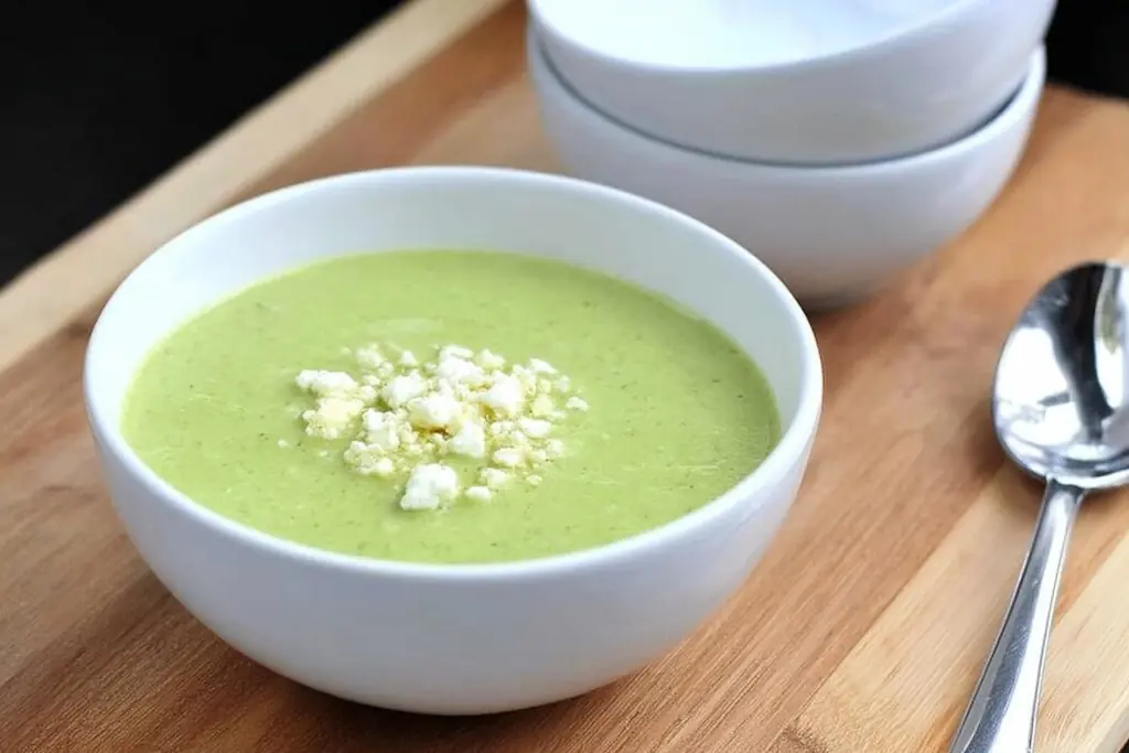 Vibrant green zucchini soup in white bowl garnished with crumbled feta cheese on bamboo cutting board with spoon