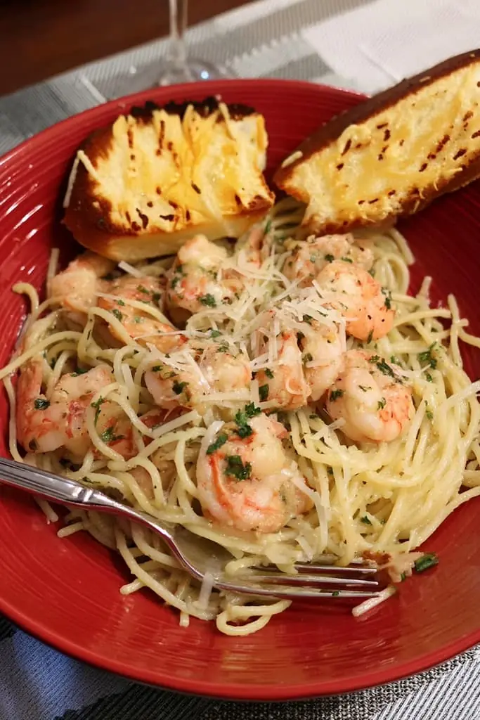 Golden garlic butter shrimp scampi served over angel hair pasta on a red plate with fresh parsley and Parmesan cheese, alongside toasted garlic bread