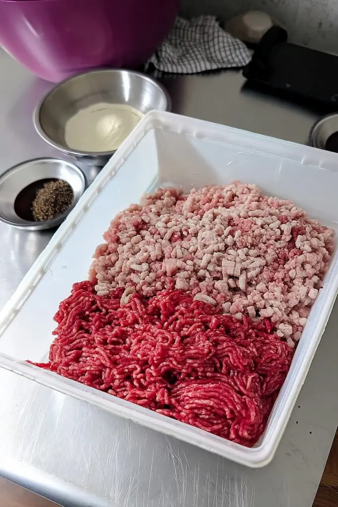 Plastic tub containing freshly ground lean beef and coarse-cut pork back fat, with small stainless steel bowls holding seasonings and curing ingredients, placed on a stainless steel countertop in a kitchen.