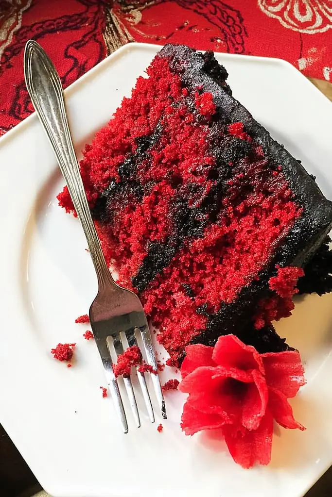 Partially eaten slice of gothic Halloween cake with red velvet layers and black buttercream on white plate with fork and red flower
