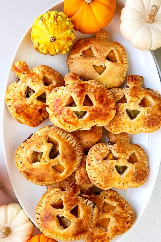 White oval platter filled with golden jack-o-lantern shaped hand pies with carved faces, surrounded by mini pumpkins