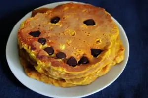 Stack of golden pumpkin pancakes with chocolate chip jack-o-lantern faces on white plate against dark blue background