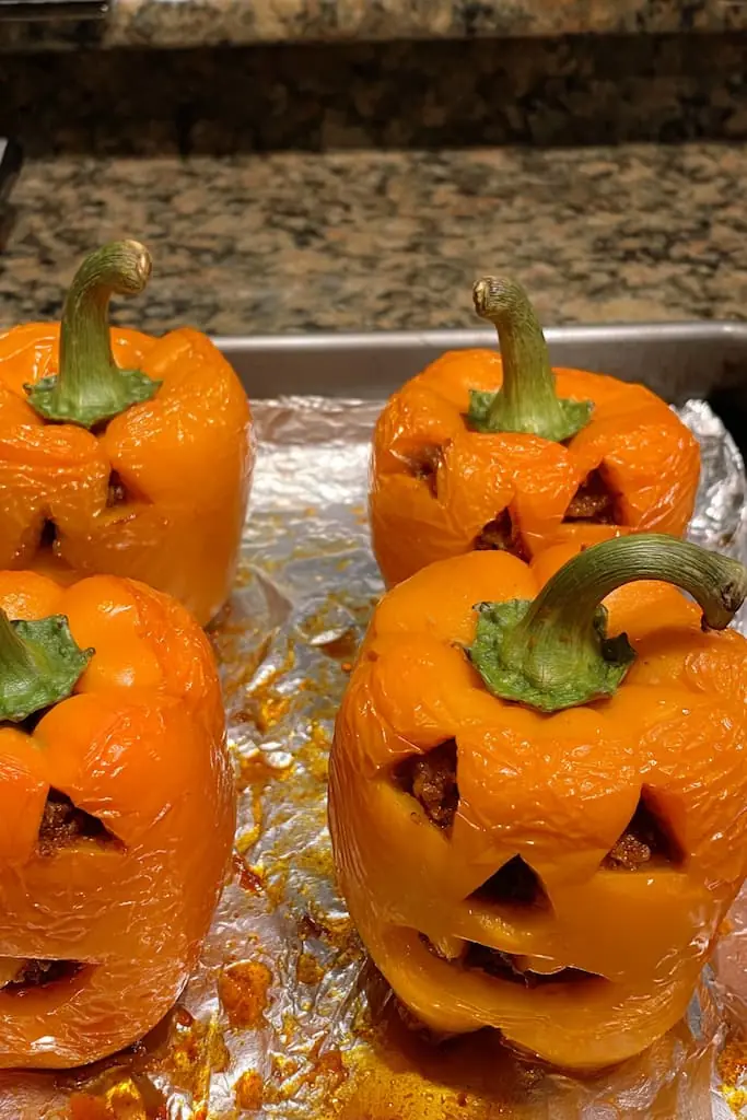 Four baked jack-o-lantern stuffed bell peppers with carved triangle faces on foil-lined baking sheet showing glossy tender peppers and beef filling