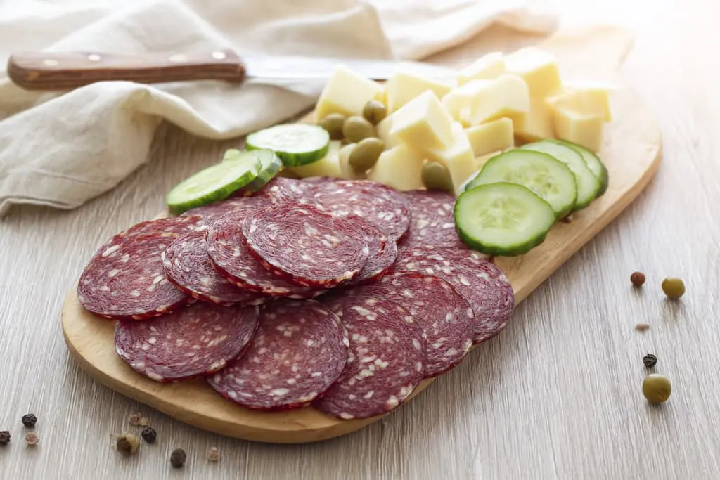 Sliced hard salami arranged on wooden cutting board with white cheddar cheese cubes, green olives, and cucumber slices for keto snacking
