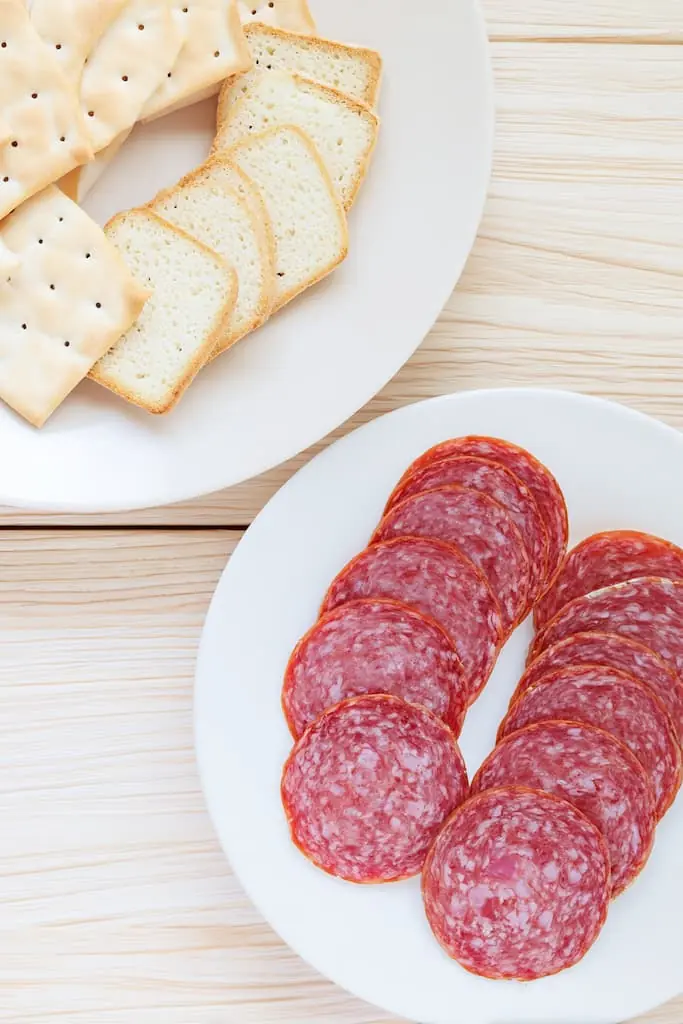 Sliced hard salami arranged on white plate next to white bread slices on separate plate, both on light wooden surface