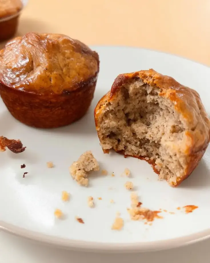 Golden brown high protein banana muffins on white plate, one broken open showing soft interior texture