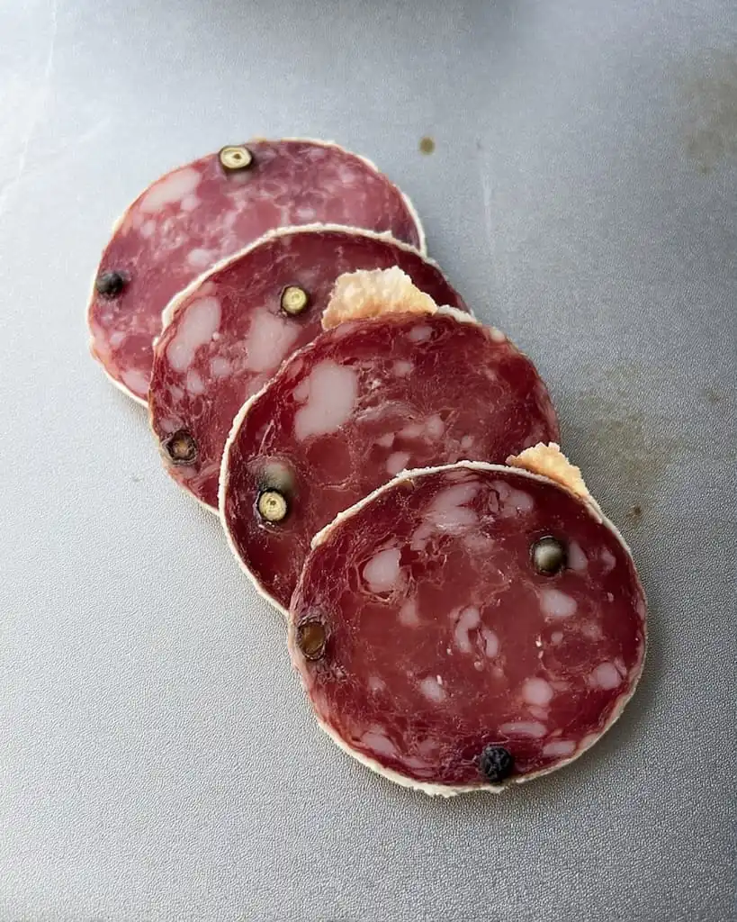 Four slices of homemade Genoa salami showing marbled pork and beef with visible whole black peppercorns and white mold rind on gray surface