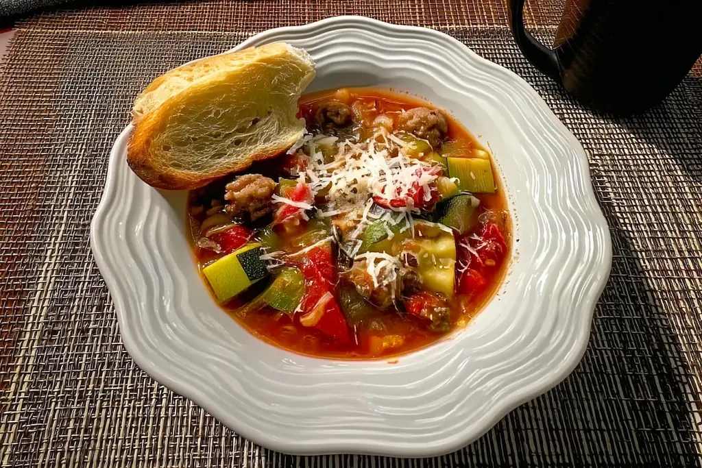 Bowl of Italian sausage zucchini soup with chunks of sausage, zucchini, tomatoes, and bell peppers topped with grated Parmesan cheese and served with crusty bread