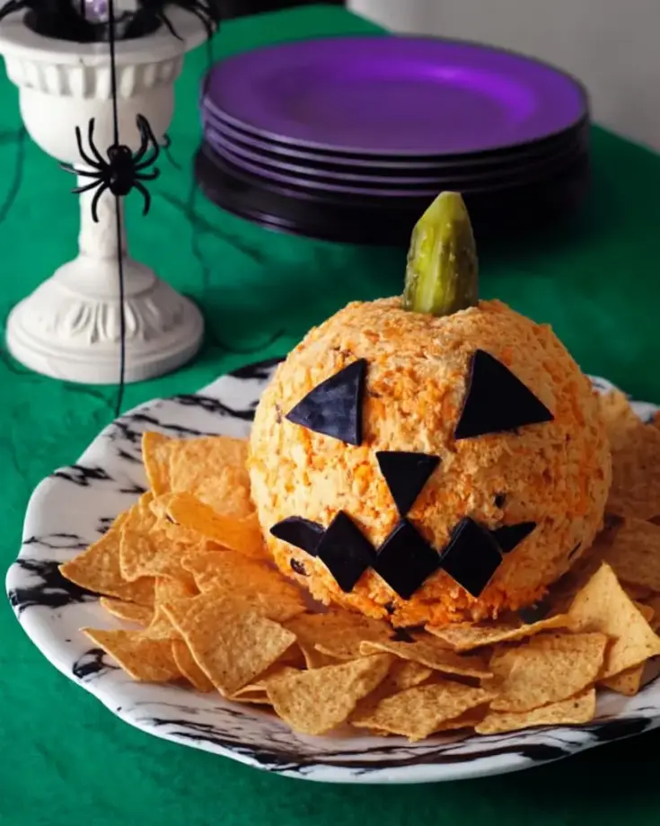 Orange Jack-o'-lantern cheese ball with black olive face and pickle stem surrounded by tortilla chips on Halloween party table