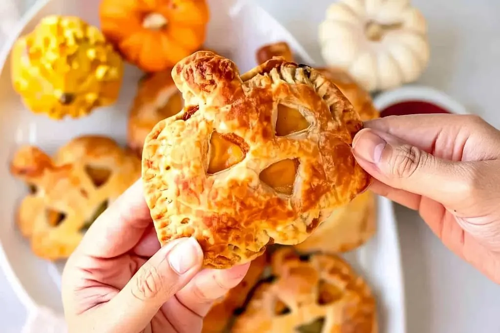 Hands holding golden jack-o-lantern hand pie showing flaky pastry layers and cheeseburger filling through carved face
