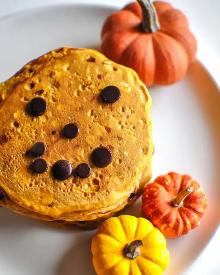 Golden pumpkin pancake with chocolate chip jack-o-lantern face on white plate surrounded by small decorative pumpkins