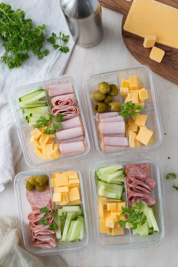 Four glass meal prep containers filled with keto-friendly deli meats including turkey roll-ups, salami, and ham paired with cheddar cheese cubes, cucumber sticks, and green olives on white marble surface